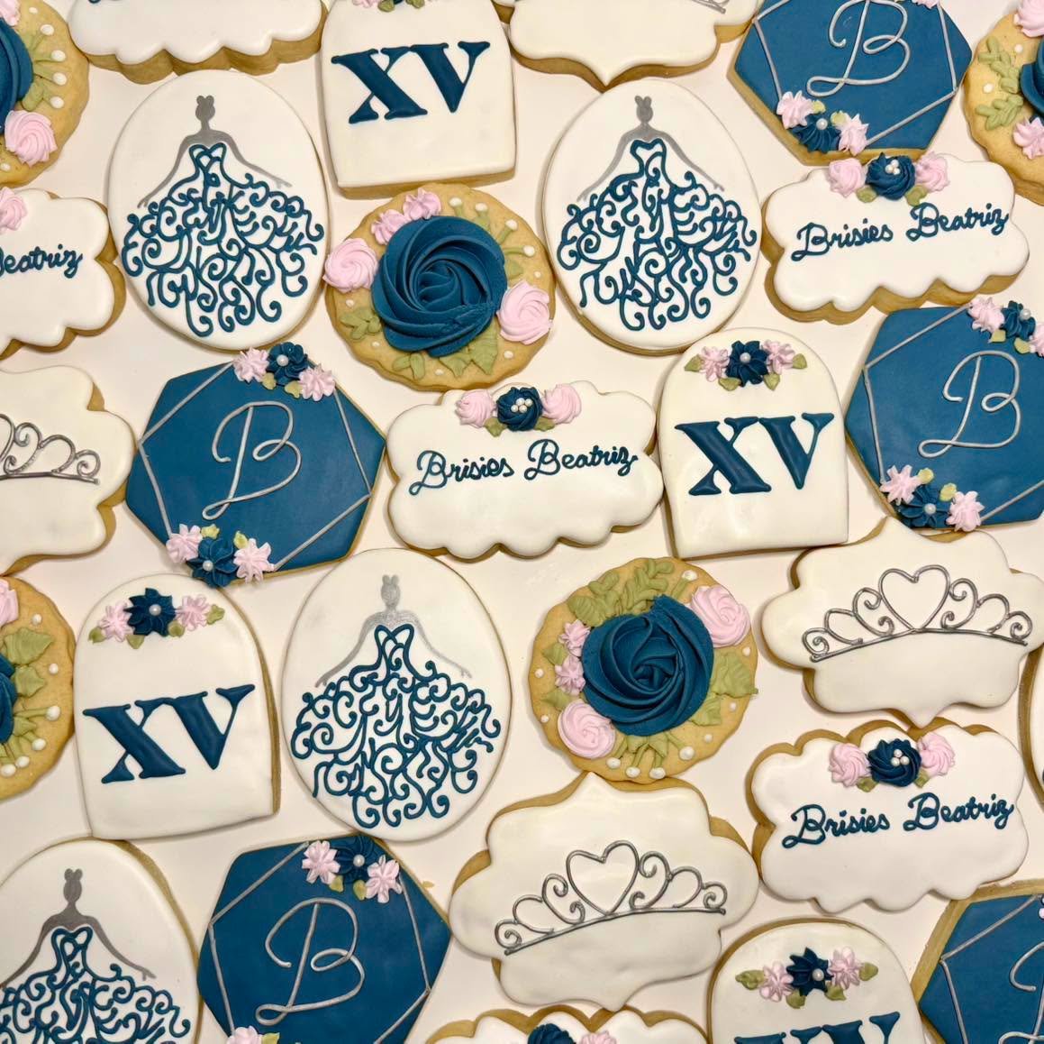 A custom set of navy and white decorated sugar cookies for a quinceañera, with XV plaques, ball gown silhouettes, tiaras, piped roses, and monogrammed hexagons.