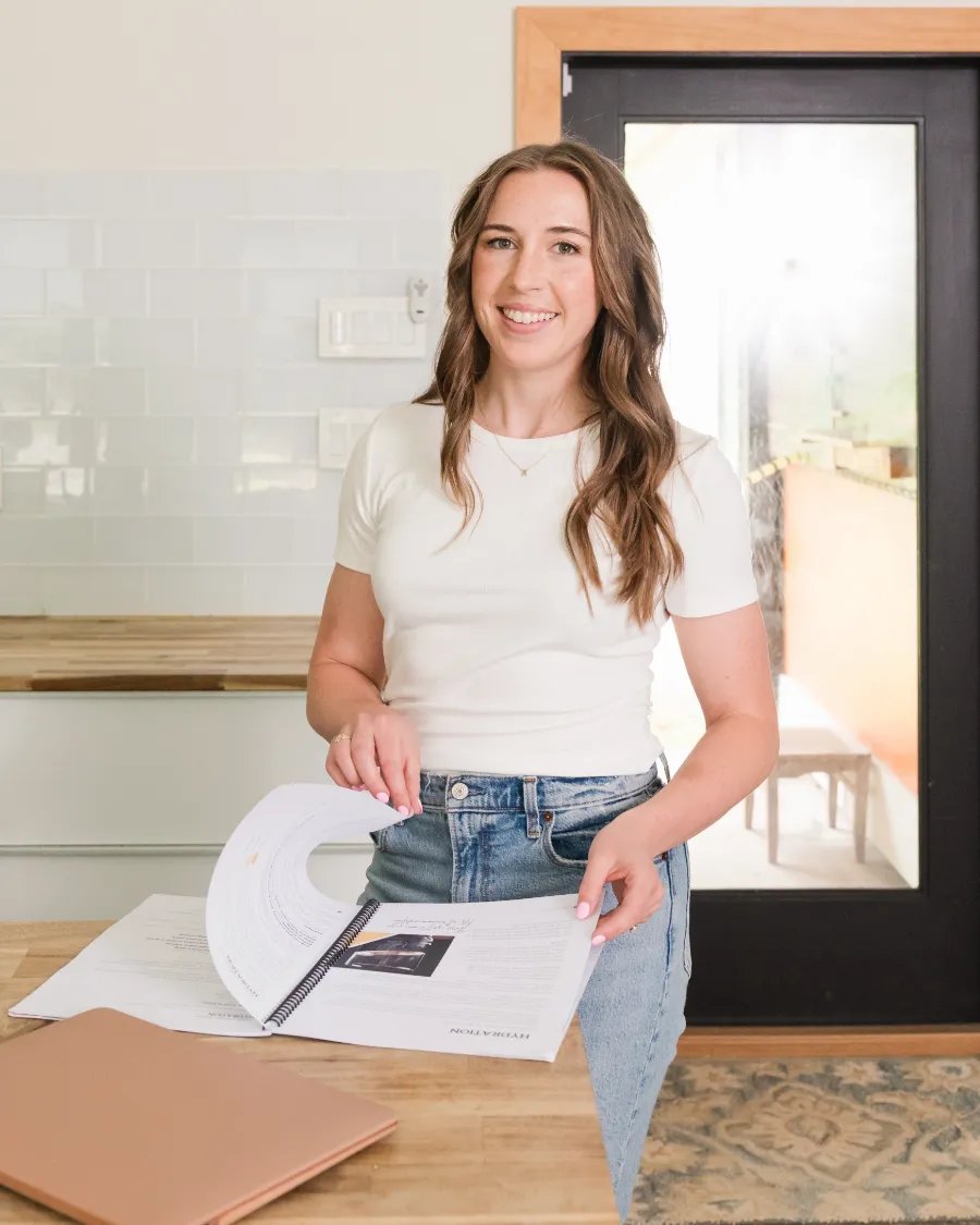 Stephanie Militano smiling as she turns the pages of a spiral-bound nutrition guide open on a wooden counter, with a laptop beside it.