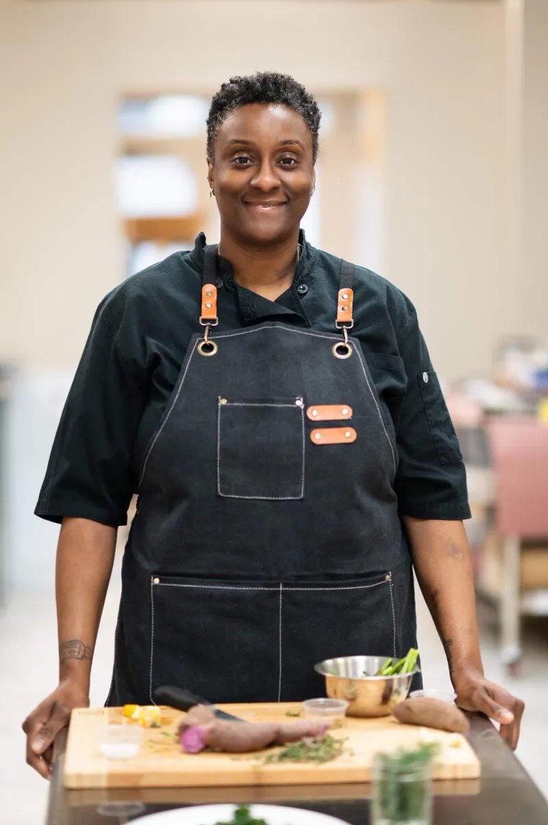 Sophia Shepherd in a chef's coat and denim apron, standing behind a cutting board of prepped herbs and vegetables in her teaching kitchen.